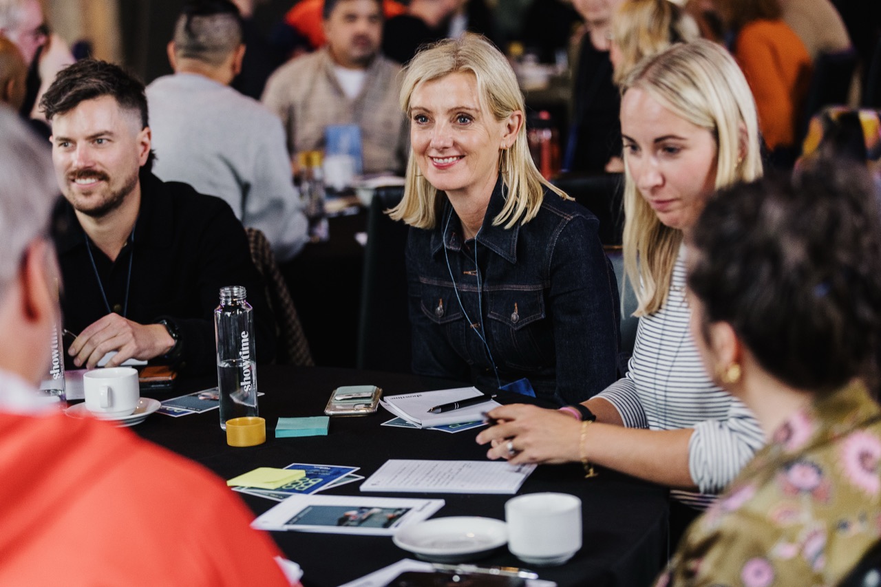 A group of people sitting at a table during a conference, with a blonde woman in a denim jacket smiling in the centre