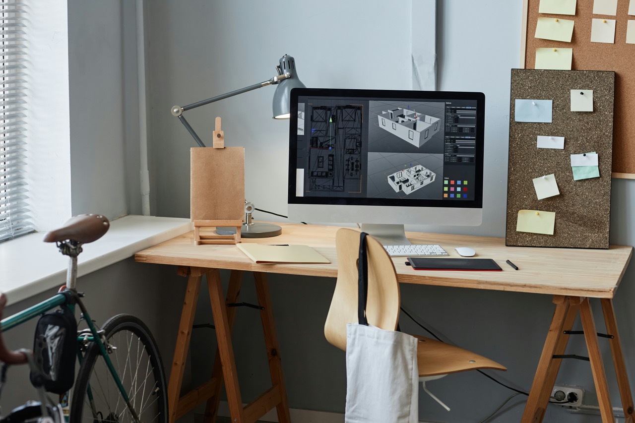 A modern workspace featuring a wooden desk with a computer displaying architectural designs, a desk lamp, a small easel, and a corkboard with notes. A bicycle is parked nearby.