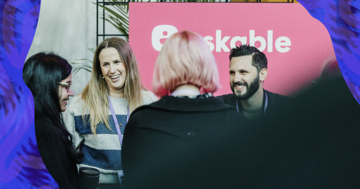 A group of four people engaged in a cheerful conversation at a social event. In the foreground, a person with pink hair is seen from behind, facing the other three. The woman in the middle has long hair and is laughing, dressed in a grey striped sweater. To her right, a woman with glasses is smiling, and to her left, a bearded man in a black shirt is also smiling. In the background, a vibrant pink banner with the text “askable” suggests a corporate or promotional setting. The group appears relaxed and happy